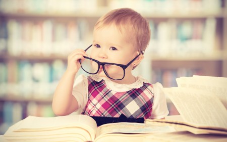 Funny Baby Girl In Glasses Reading A Book In A Library
